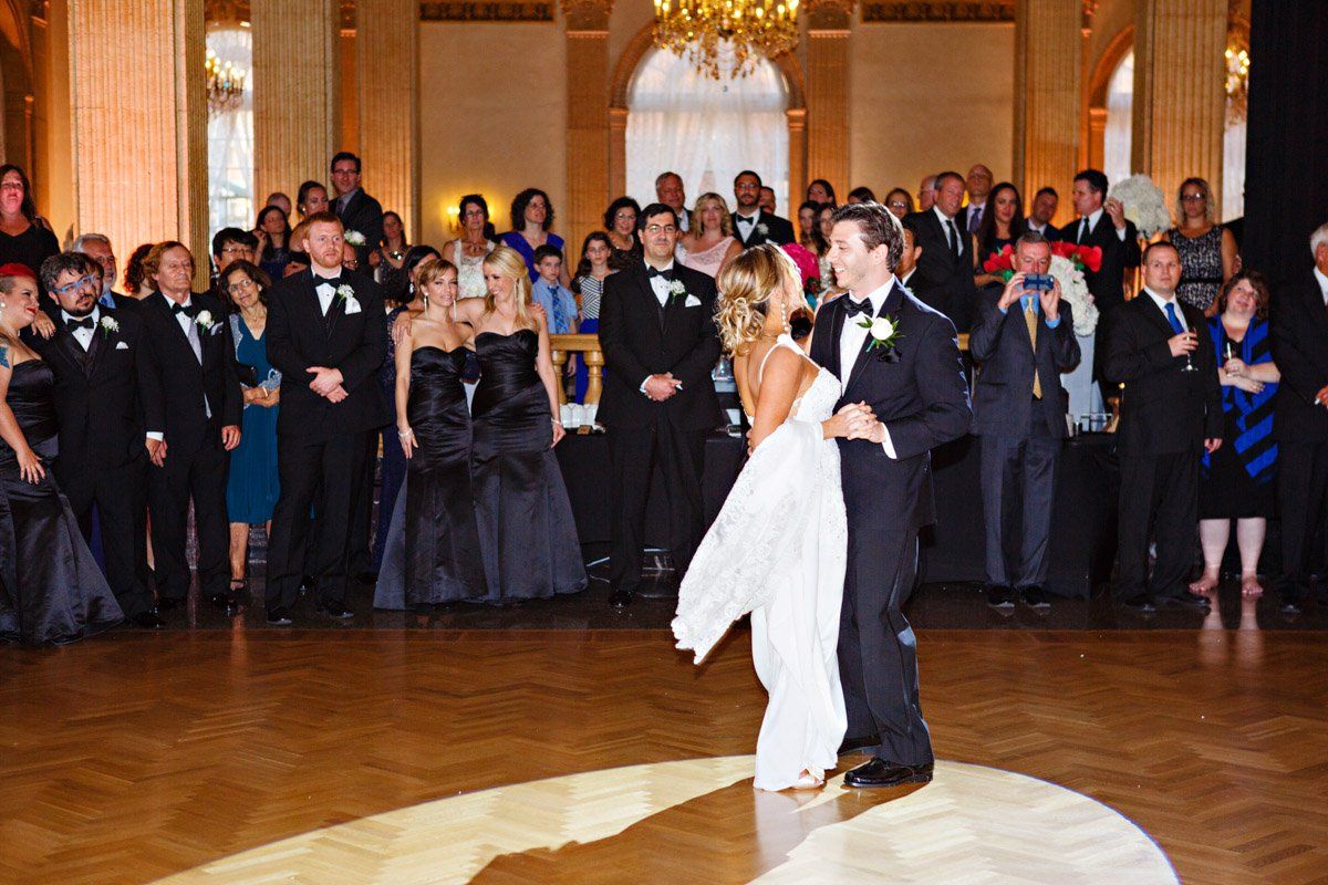 Newlyweds dance as guests watch at wedding reception. Formal attire, ballroom setting, golden light.