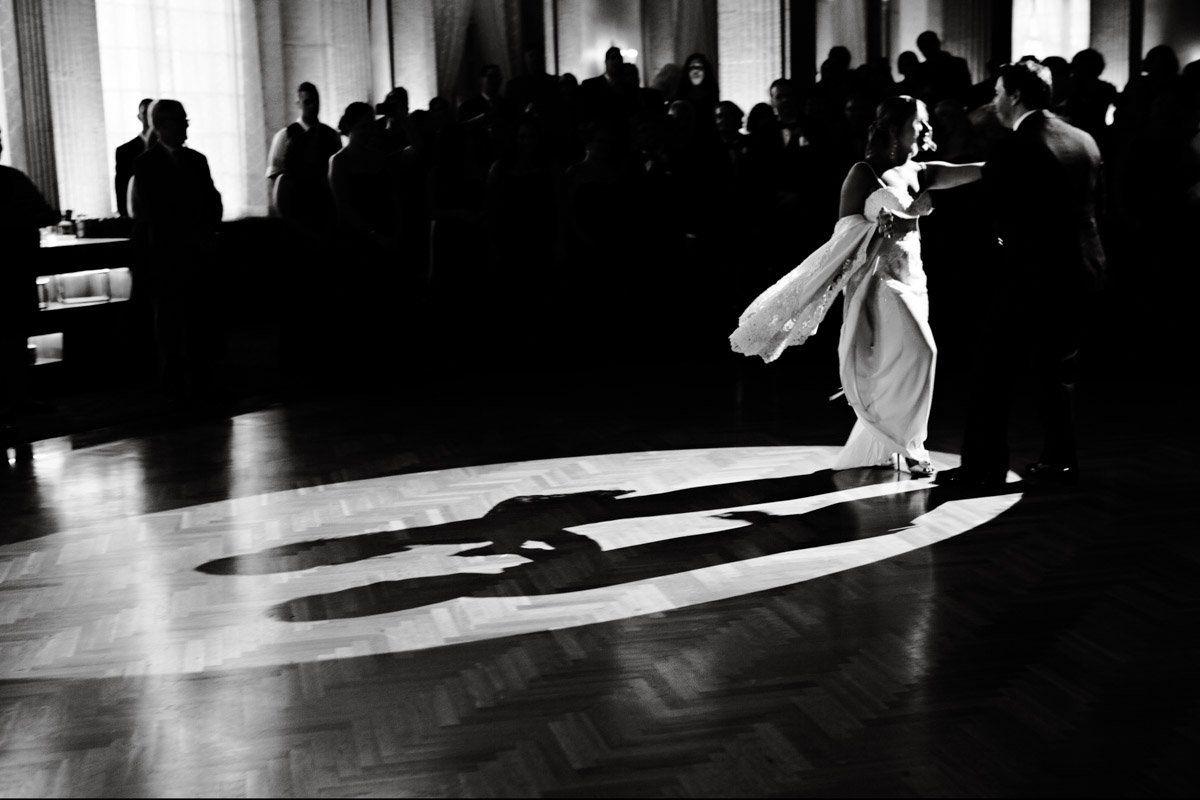 Couple dancing in spotlight on a wood floor at a formal event, shadows cast.