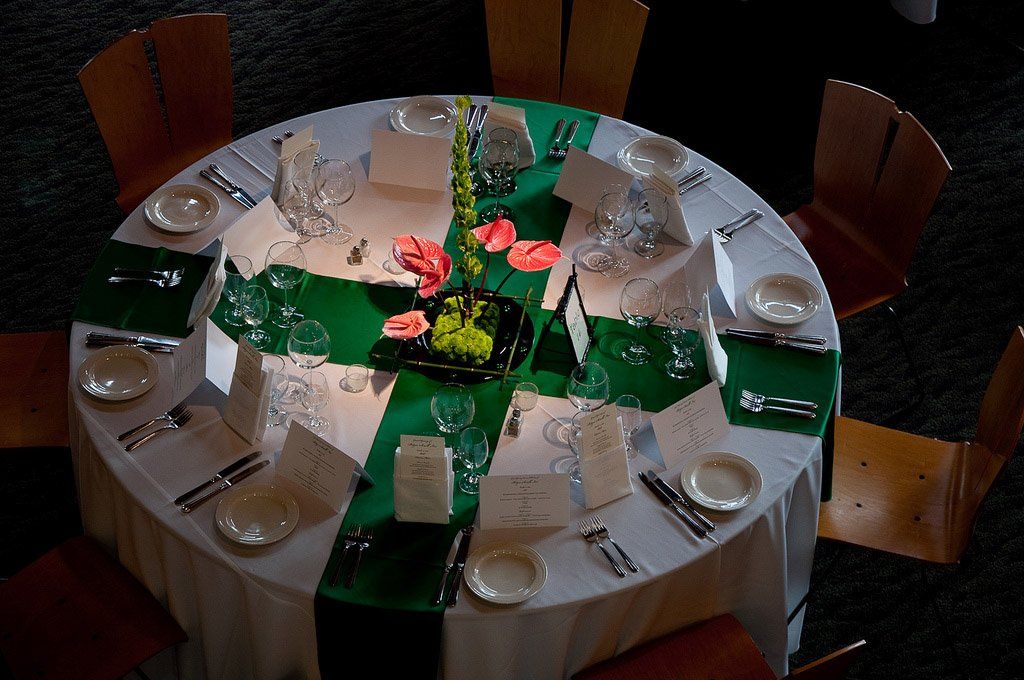 Round table set for a formal event, with green accents and floral centerpiece, surrounded by wooden chairs.