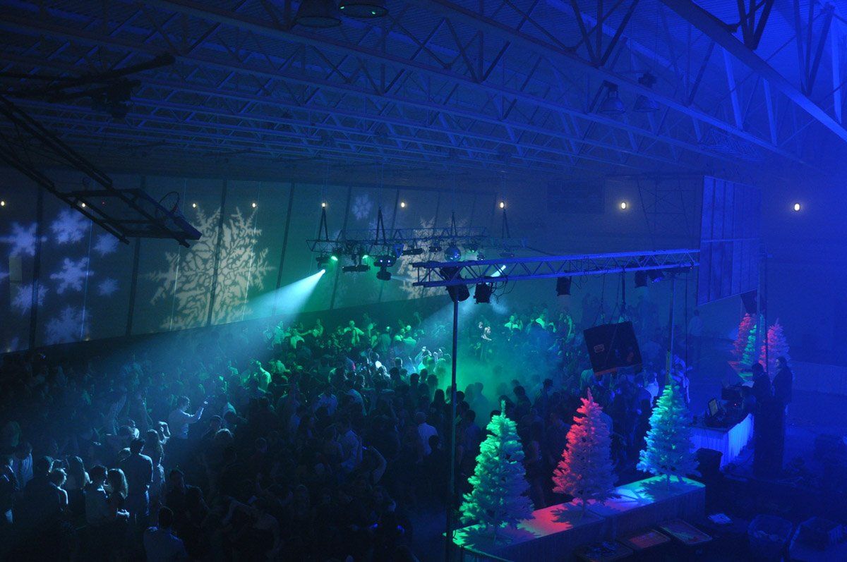 Large indoor party with DJ, green, blue, and white lighting, crowd, Christmas trees, and projected snowflakes.