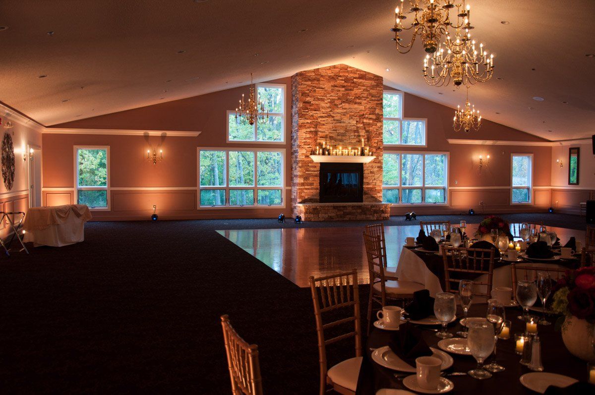 An empty event space with a fireplace, large windows, chandeliers, and tables set for a gathering.
