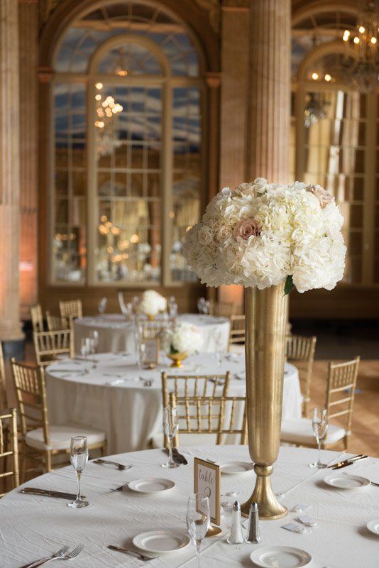 Elegant wedding reception table with tall gold centerpiece, white linens, and chairs in a grand hall.