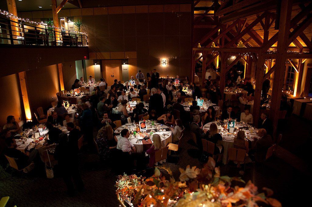 Wedding reception in large hall, round tables, many guests seated, warm lighting.