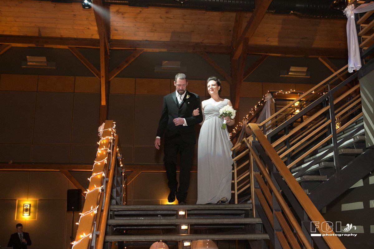 Father escorts bride down a stairwell at a wedding. The bride wears white, carries a bouquet. Venue is rustic.