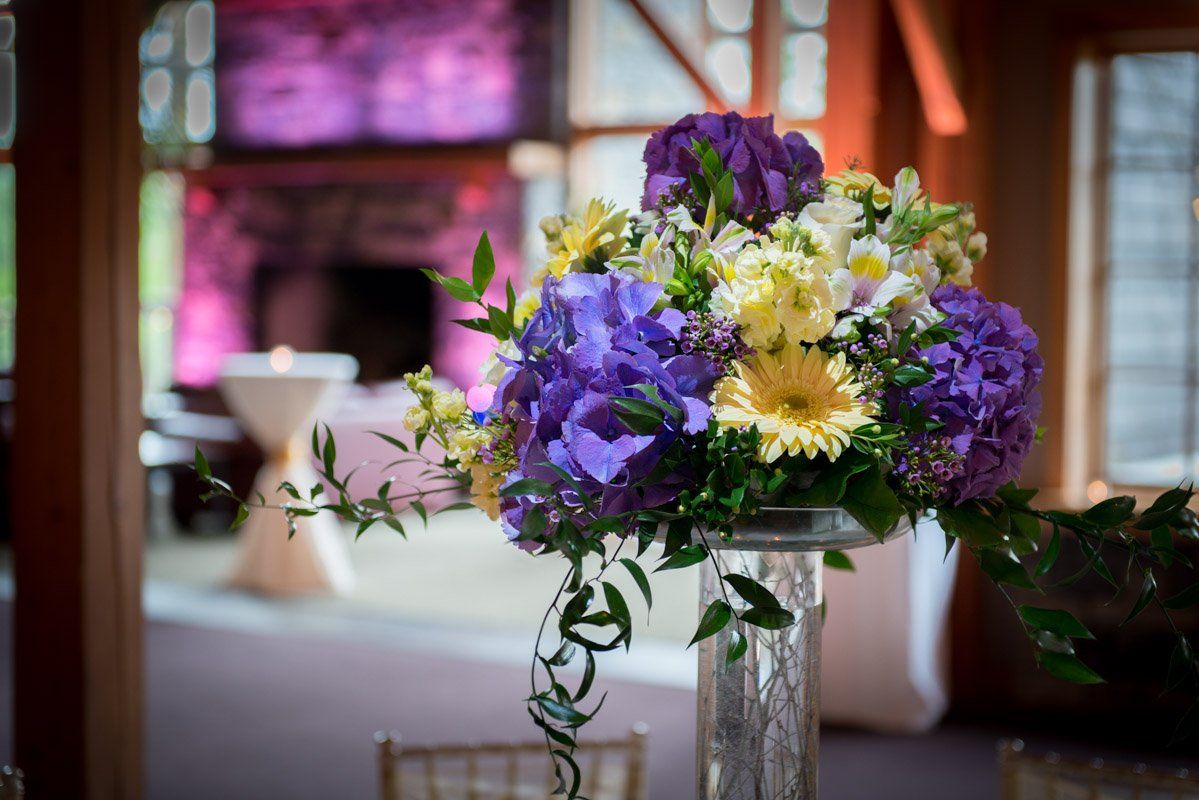 Purple and yellow floral arrangement on a tall glass vase, indoors at an event.