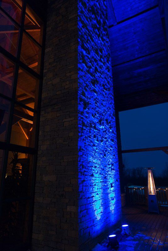 Blue spotlights illuminate a tall brick pillar outside at dusk. Orange light glows from a nearby window.