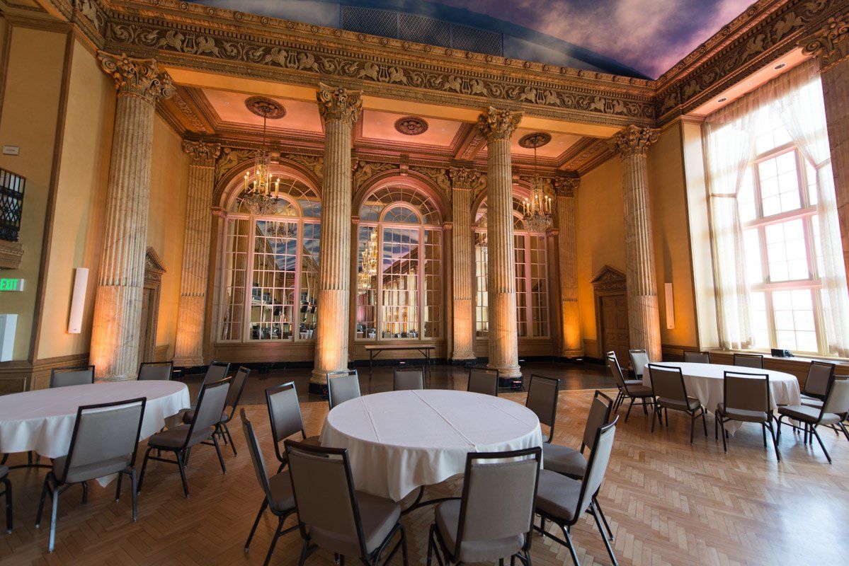 Elegant event space with round tables, chairs, ornate pillars, and large windows with sunlight.