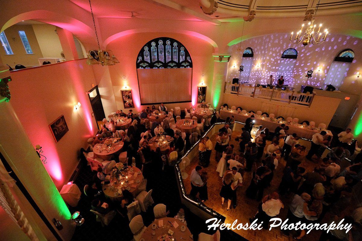 Wedding reception in a large hall with round tables, people, and colorful uplighting.