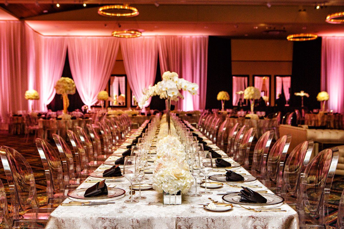 Long banquet table set for an event with clear chairs, white flowers, and pink lighting.