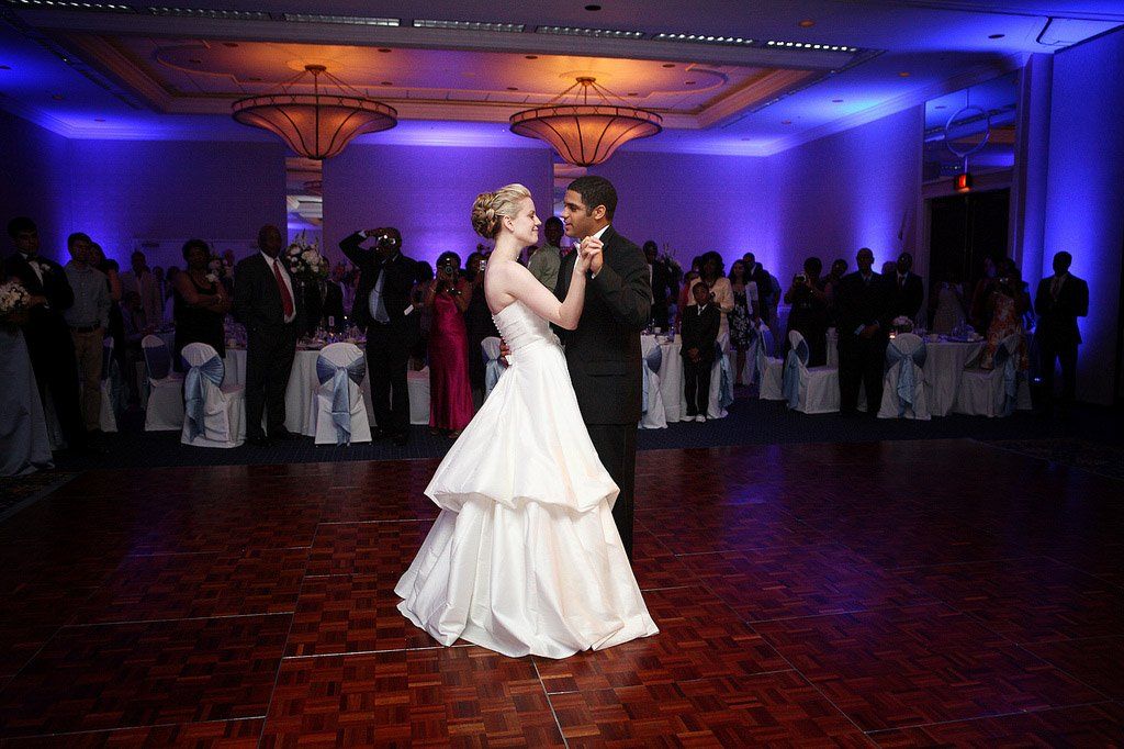Bride and groom dance at wedding reception; guests watch, blue lighting, ballroom.