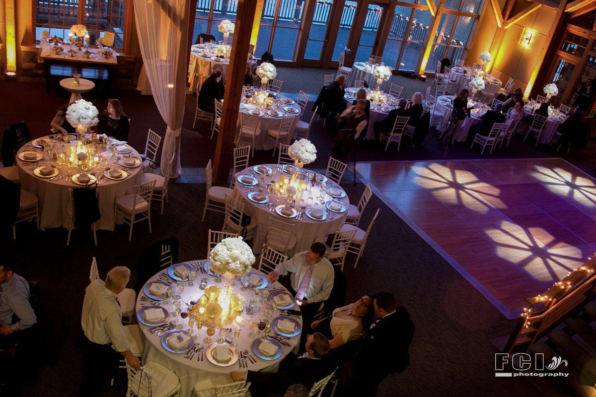 Wedding reception: round tables with white linens, floral centerpieces, guests mingling; wooden dance floor with flower light projections.
