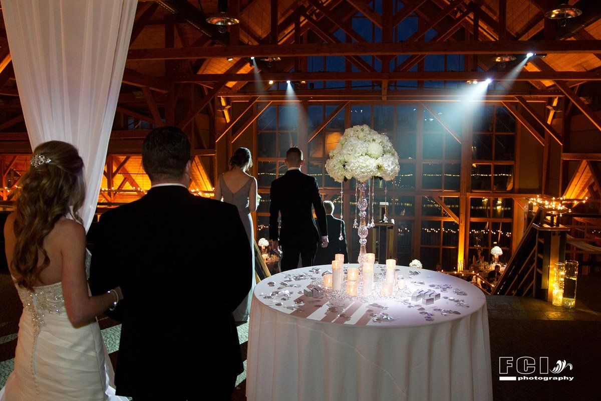 Bride and groom at wedding ceremony, with tall flower arrangement and candles on table. Wooden interior setting.