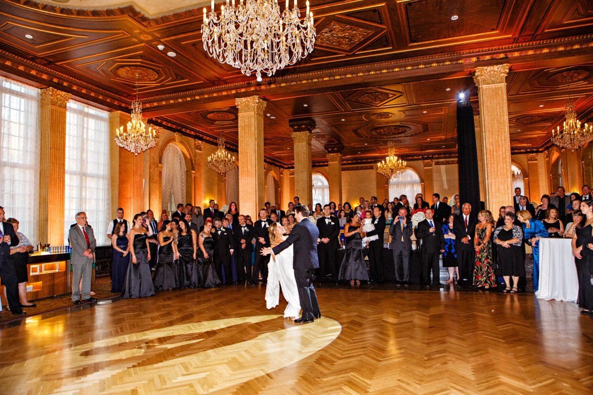Couple dancing at a wedding reception. Guests watch in a grand ballroom with chandeliers and columns.