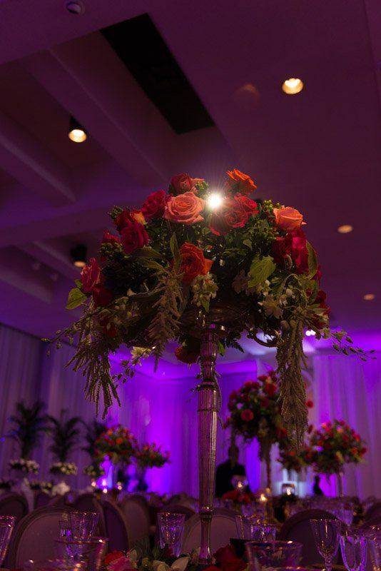Floral centerpiece on a tall stand, with red and orange roses, set on a table in a room with purple lighting.