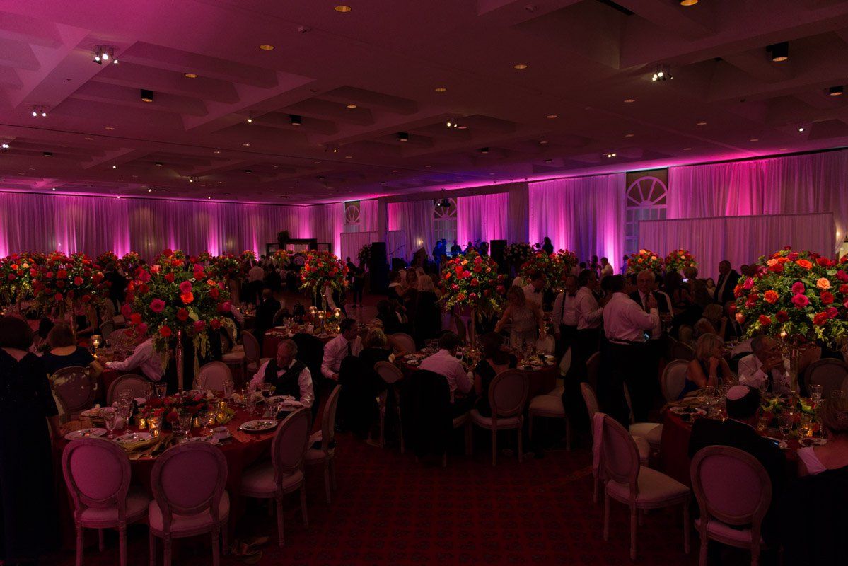Formal event in a large hall with tables, floral centerpieces, and pink lighting. People are seated, socializing.