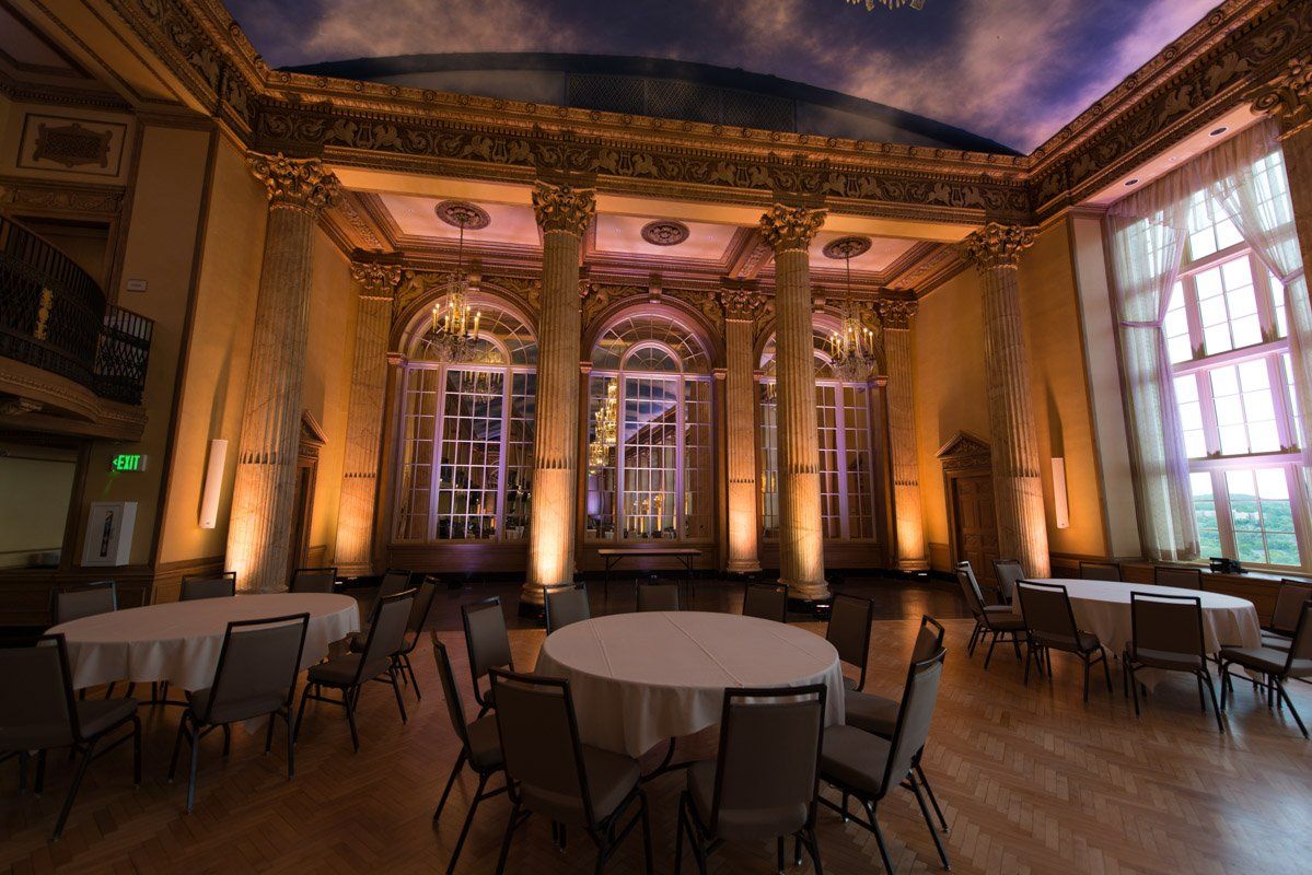 A large, ornate ballroom with round tables and chairs. Windows and columns with warm lighting.