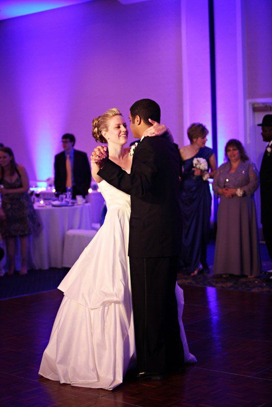 Bride and groom dance at a wedding reception; purple-lit ballroom, smiling, she wears white gown, he wears black suit.