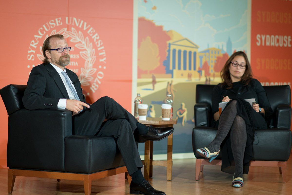 Man and woman seated on stage, in front of a Syracuse University backdrop. They appear to be at a speaking event.