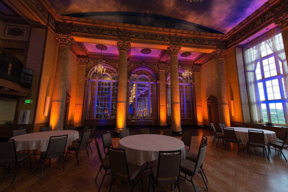 Elegant ballroom with round tables, chairs, columns, and colorful lighting, featuring orange, purple, and blue hues.