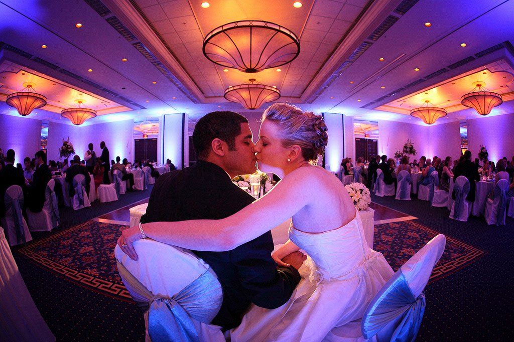 Bride and groom kissing at a wedding reception. Purple and orange lighting, crowd in the background.