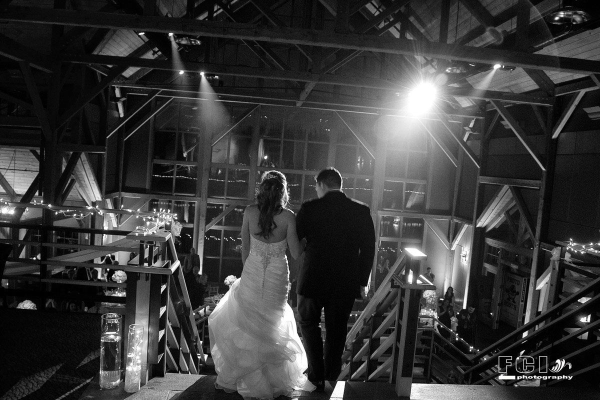 Bride and groom walking away, hand-in-hand, in a dimly lit industrial venue; black and white photo.