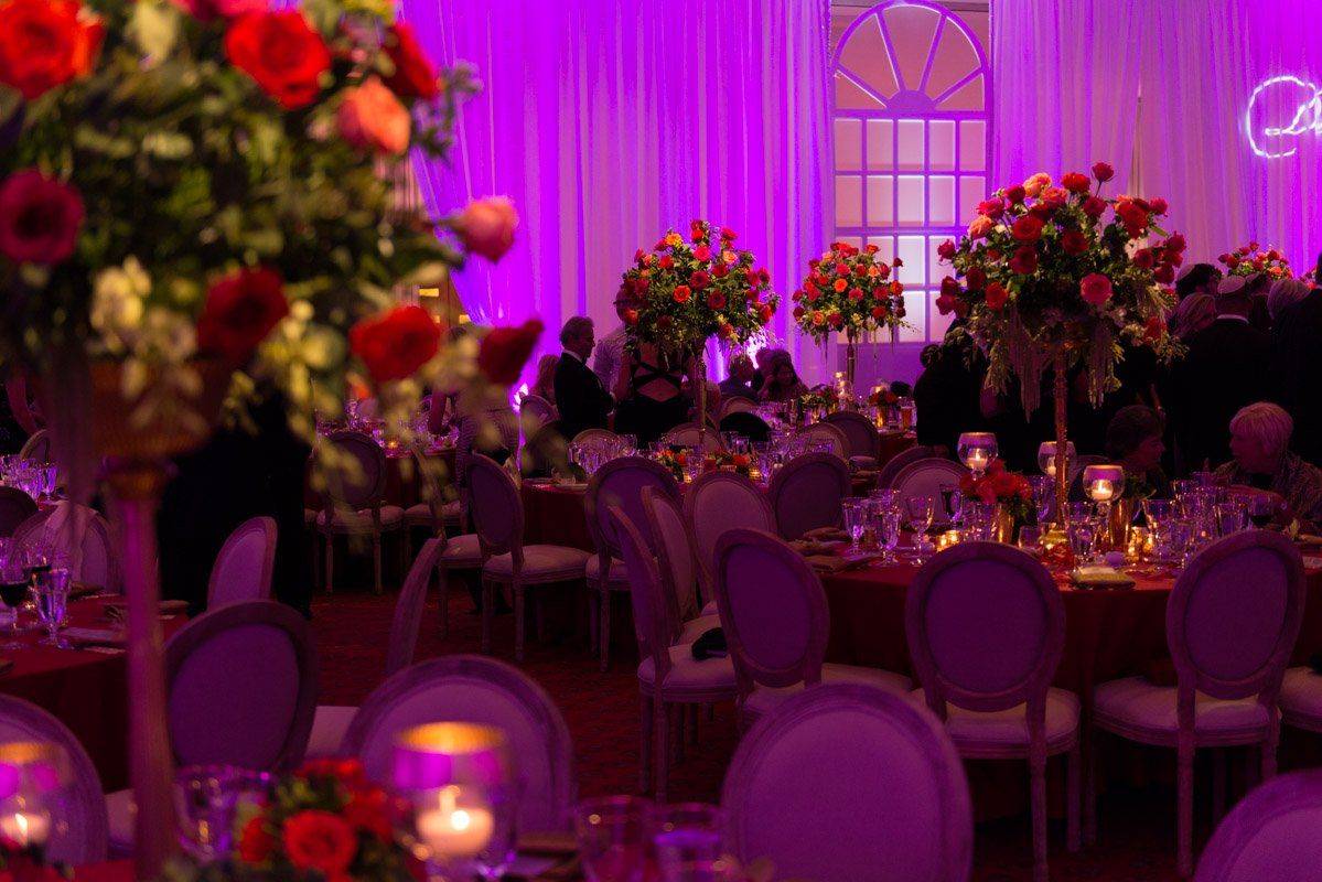 Formal event with tables, chairs, and tall floral centerpieces. Purple lighting and red table cloths.