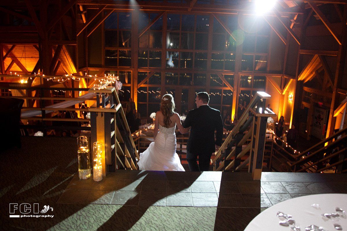 Bride and groom walk down wooden stairs during wedding reception, warm lighting, indoor barn setting.