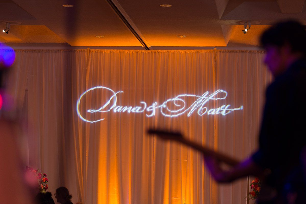 Dana & Matt name projection on a fabric backdrop, illuminated with orange light; a musician plays guitar in the foreground.