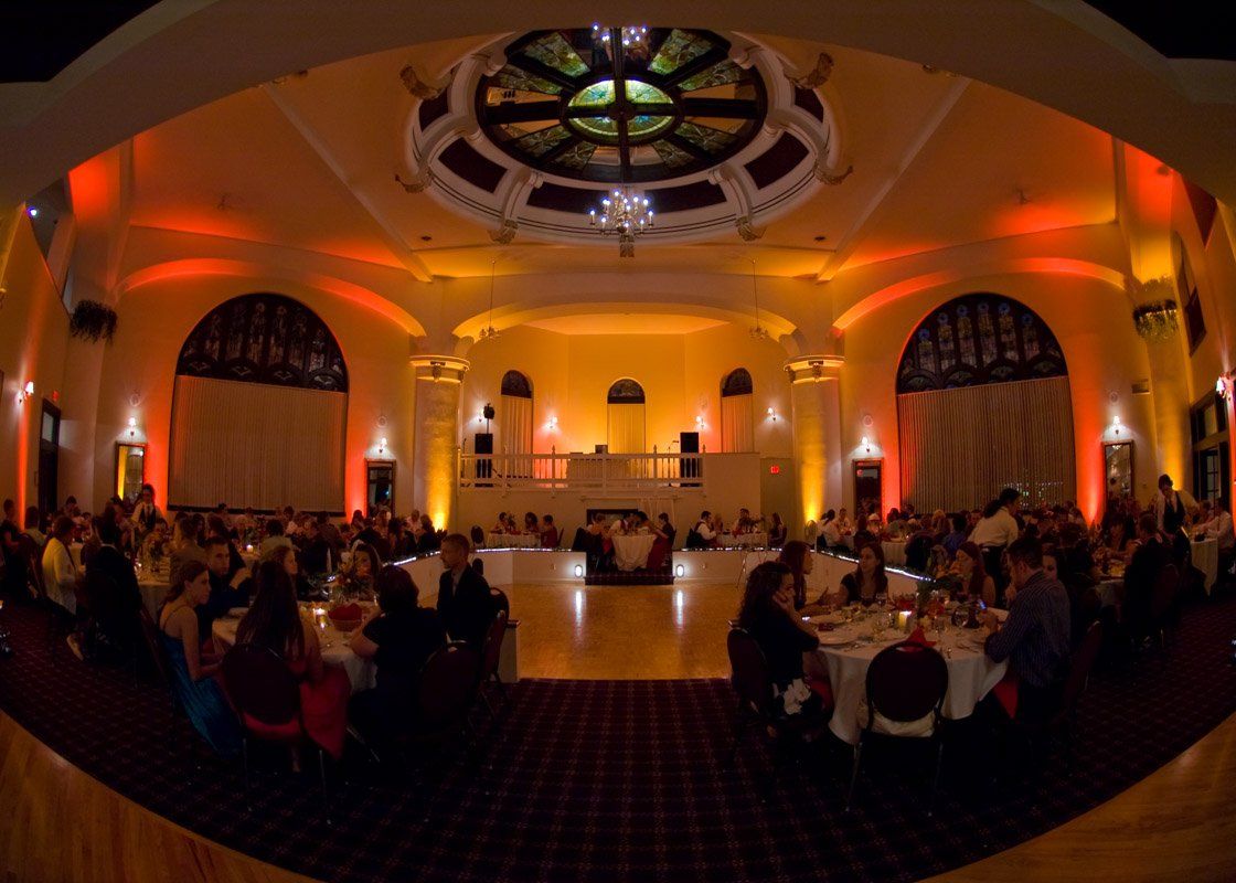 Ballroom with people at tables, lit with orange and yellow lights, and a stained-glass ceiling.