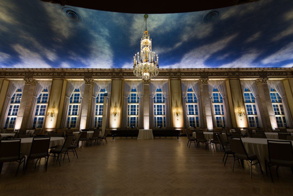 Elegant ballroom with ornate details, windows, a large chandelier, and tables set for a gathering.
