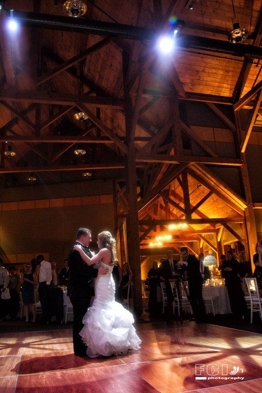 Bride and groom dancing in a barn with wooden beams, soft lighting, and guests in the background.