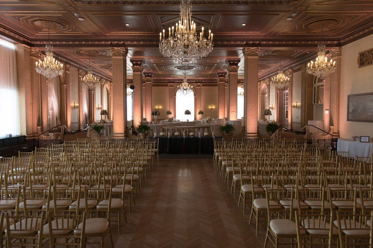 Elegant ballroom set for a wedding ceremony, rows of gold chairs face a stage, lit by chandeliers.
