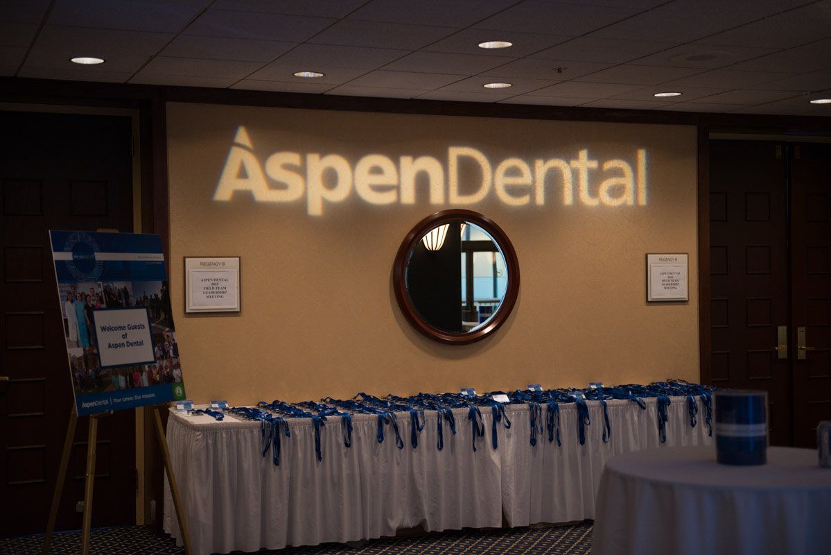 Aspen Dental logo projected on wall above a table with name tags. Dark room, brown and white decor.