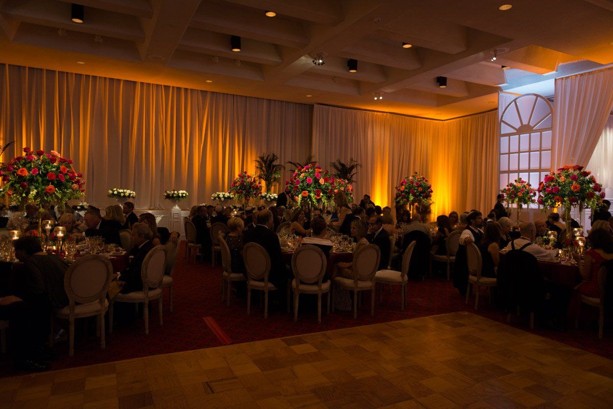 Elegant ballroom with tables, guests, flowers, and warm lighting.