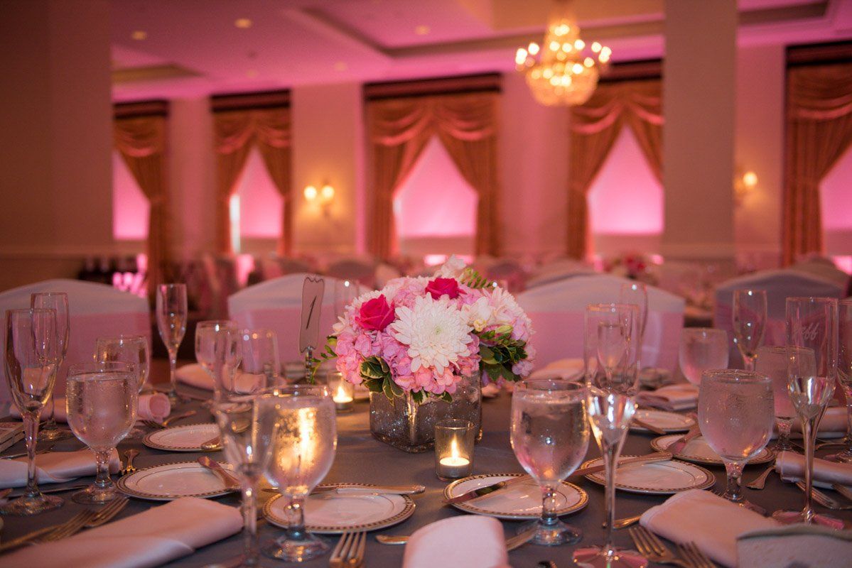 Elegant wedding reception table set with flowers, candles, and glassware under pink lighting.