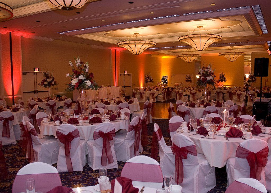 Wedding reception in a large ballroom with round tables, white linens, red accents, and ornate chandeliers.