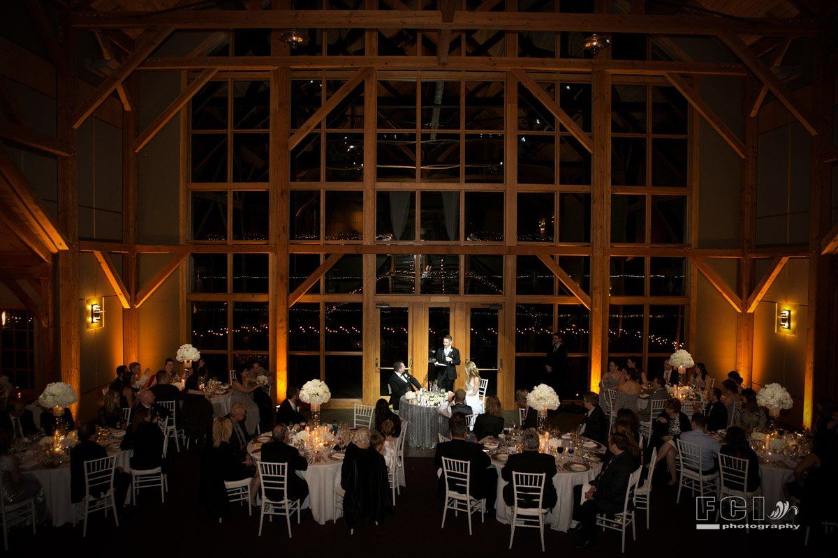 Wedding reception in a large hall with large windows; guests at tables, couple at a table in front of windows.