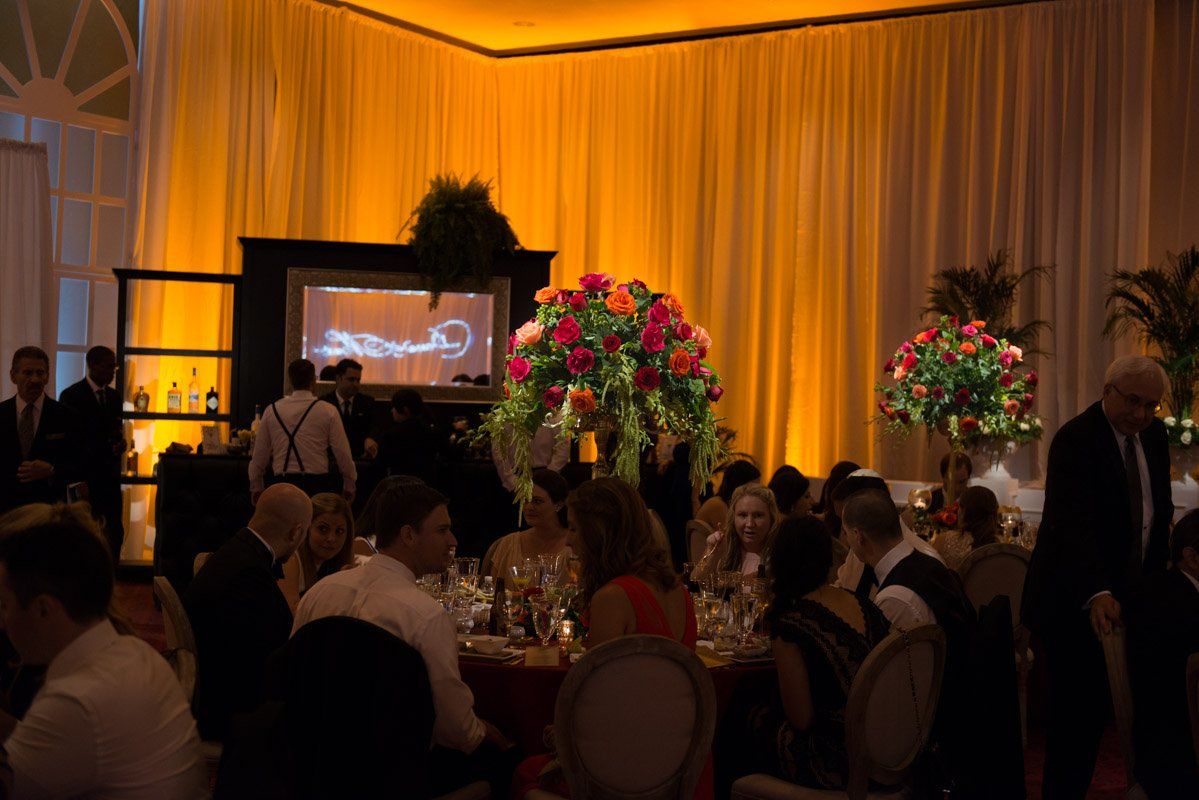 Elegant event with guests seated at tables, large floral arrangements, and draped orange lighting.