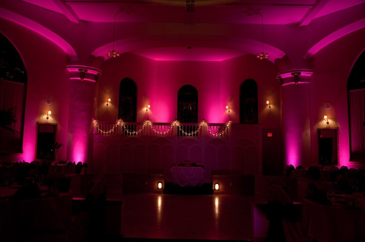 Pink uplighting illuminates an empty hall with arches, a balcony, and stage.