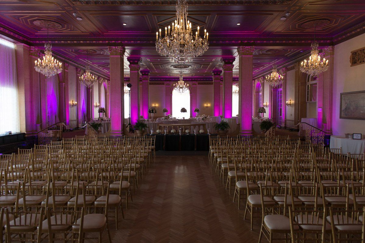 Elegant ballroom set up for an event with rows of gold chairs, chandeliers, and pink uplighting.