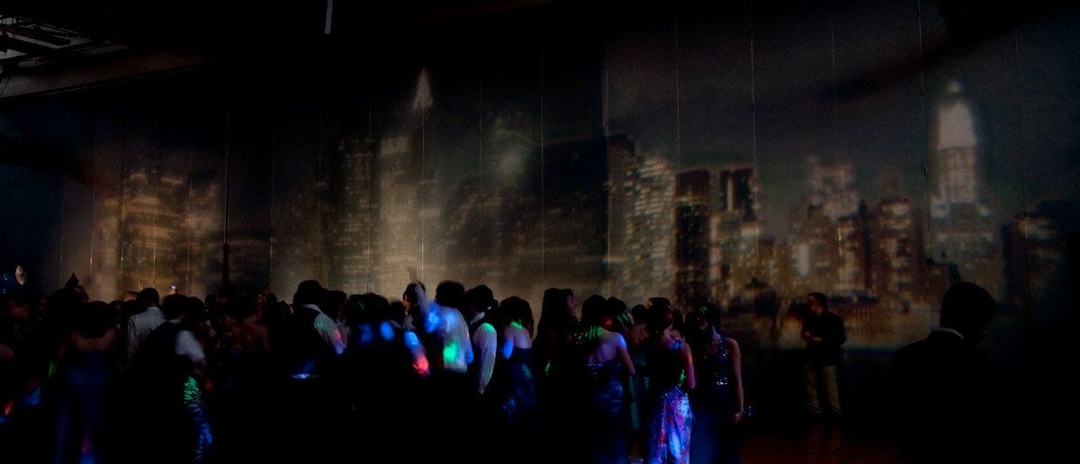 People dance in a dark room with a city projection on the wall. Blue and green lights illuminate figures.