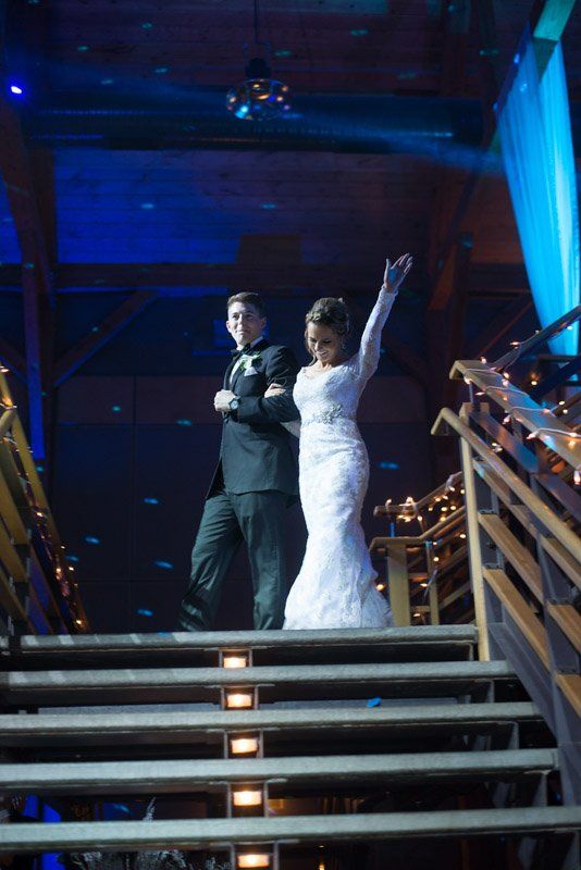 Couple in formal attire, descending illuminated stairs at a dance, woman waving.