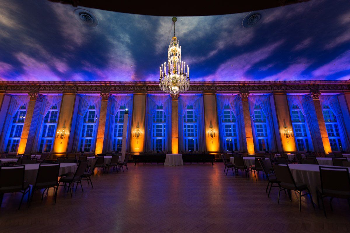 Elegant ballroom with blue and orange uplighting, chandelier, and tables set for a formal event.