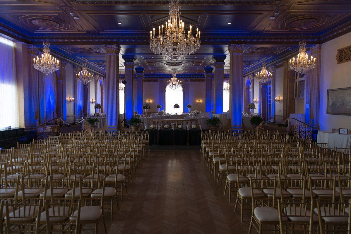Rows of gold chairs face a stage in a formal ballroom with chandeliers and blue uplighting.