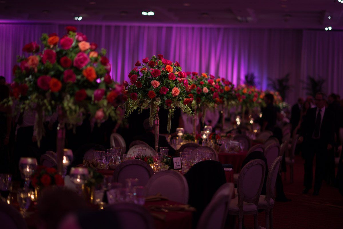Elegant banquet hall with floral centerpieces, tables, and chairs. Lit with pink and purple lights.
