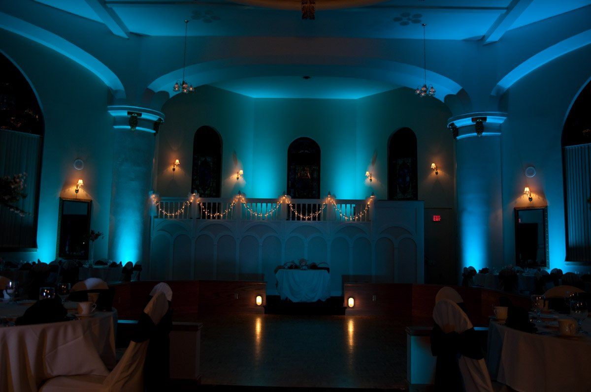 Blue-lit ballroom with stage. Tables set for event, with light reflecting on the floor and up the walls.