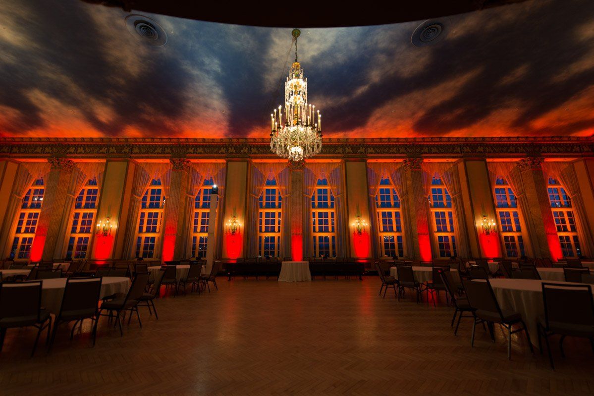 Elegant ballroom lit with red and orange hues, a chandelier hangs from a painted cloudy ceiling.