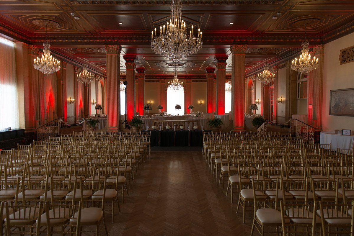 Rows of gold chairs face a stage in a grand room with red uplighting and chandeliers.