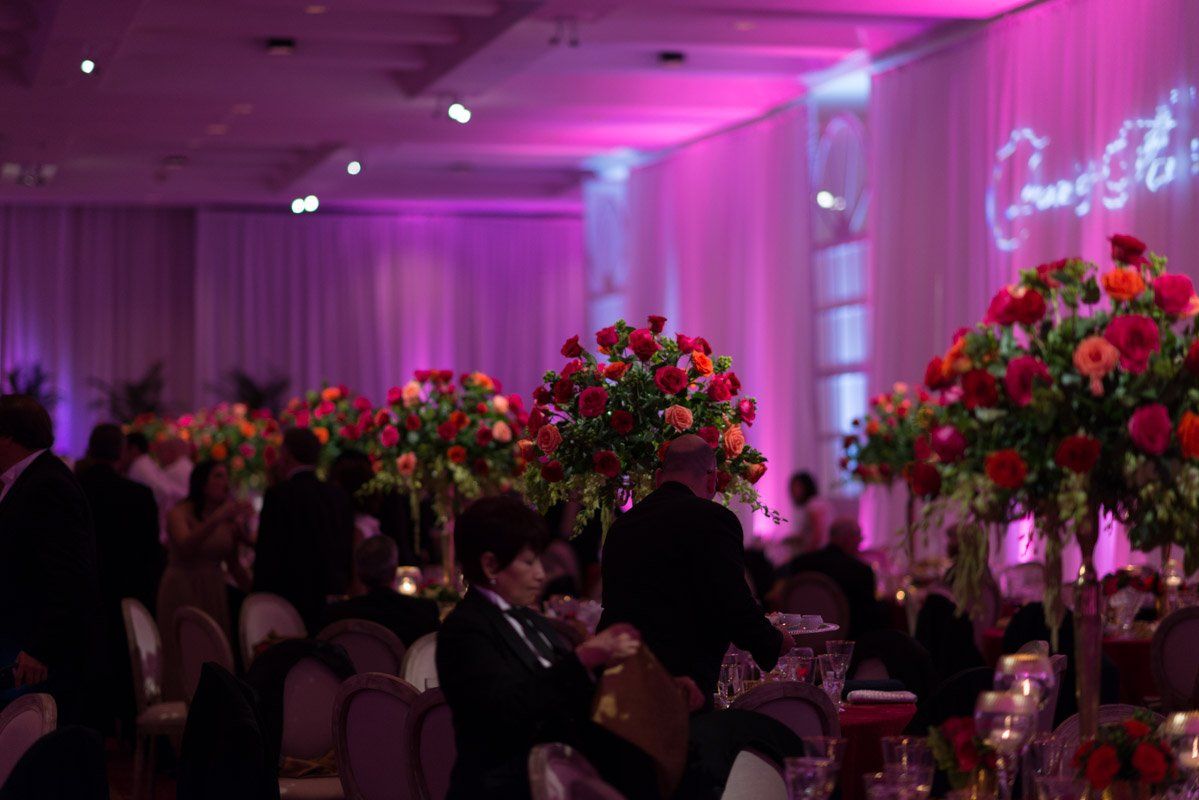 A festive event with colorful floral centerpieces on tables. Purple lighting illuminates the room.
