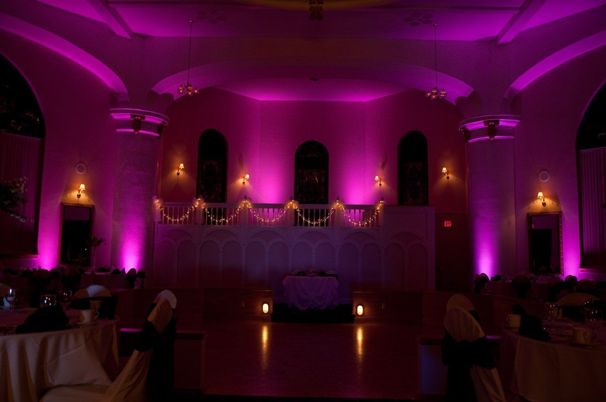 A ballroom with purple uplighting illuminates the stage and architectural details. Tables are set for a formal event.